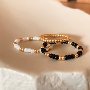 Könnte beinhalten: Drei Perlenringe auf einer weißen Keramikplatte. Ein Ring ist weiß mit goldenen Perlen, einer ist schwarz mit goldenen Perlen und einer ist komplett aus goldenen Perlen.