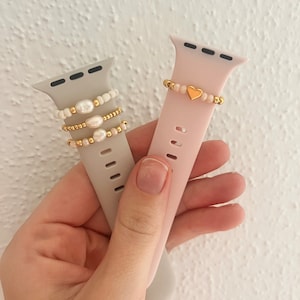 Könnte beinhalten: Zwei Smartwatch-Armbänder. Eines ist grau mit weißen Perlen und goldenen Perlen. Das andere ist rosa mit einem goldenen Herz und Perlen. Beide sind für Smartwatches.