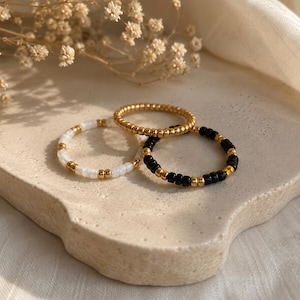 Könnte beinhalten: Drei Perlenringe, die zusammen angeordnet sind. Ein Ring besteht aus kleinen metallisch goldenen Perlen. Ein zweiter Ring zeigt abwechselnd weiße und goldene Perlen. Der dritte Ring besteht aus abwechselnd schwarzen und goldenen Perlen.