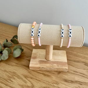 Könnte beinhalten: Drei Perlenarmbänder mit den Buchstaben "PMS" auf jedem Armband. Die Armbänder werden auf einem Holzständer präsentiert. Die Armbänder sind rosa, pfirsichfarben und weiß.