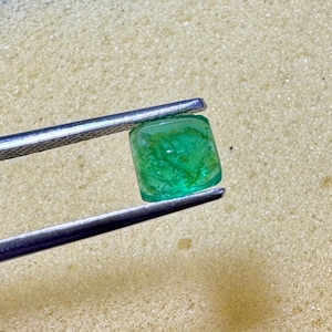 May include: A vivid green gemstone, held by silver tweezers. The gem is square with rounded edges and internal patterns. The background is a light tan, textured surface, contrasting with the jewel's colour and clarity.