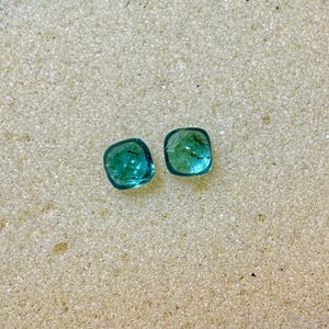 May include: Two square-cut, teal-colored gemstones with rounded corners are displayed. The gems have a translucent quality, with visible internal features. They are set against a textured, off-white surface, creating a contrast that highlights their color and shape. These gems could be used for jewelry.