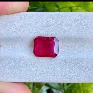 May include: A rectangular-cut ruby gemstone in a vivid red colour. The gem is displayed on a white, textured surface, with a small white ribbon. Green foliage forms the background.