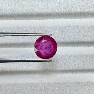 May include: A faceted, round, deep red gemstone held by silver tweezers. The gem has a polished surface and a star-like pattern visible within. The background is a textured white surface.