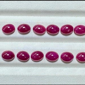 May include: Ten oval-shaped, polished ruby gemstones arranged in two rows on a white surface. The rubies are a deep, vibrant red color with a smooth, glossy finish, reflecting light.
