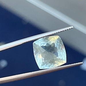 Peut inclure: Une pierre précieuse bleu pâle de coupe carrée est tenue par une pince. La pierre précieuse a une surface à facettes, reflétant la lumière. Le fond est un dégradé de bleu et de gris.