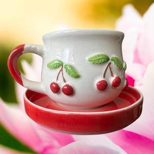 May include: A ceramic teacup and saucer set. The cup is off-white with red cherries and green leaves. The handle and the rim of the saucer are red. The saucer has a red and white striped pattern.