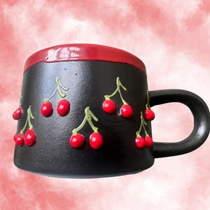 May include: Black ceramic mug with a red rim and handle, adorned with raised red cherry designs. The cherries are attached to green stems, creating a 3D effect. The mug is set against a soft, pink and red background.