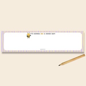 May include: A rectangular notepad with a white centre and a border featuring vertical light purple and yellow stripes. A cartoon bee illustration appears next to the text IT'S GONNA BEE A GOOD DAY! and the brand name MOMO'S PUN. A wooden pencil is included.