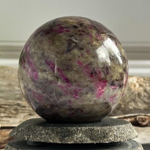 May include: A polished sphere of pink and grey ruby in zoisite, a natural gemstone, resting on a stack of grey stone coasters.