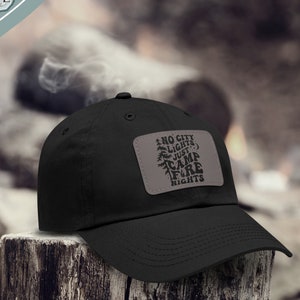 Pode incluir: Boné de beisebol preto com um remendo marrom que diz "Sem luzes da cidade, apenas noites de fogueira".