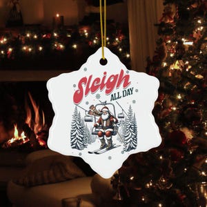 May include: White snowflake-shaped Christmas ornament featuring Santa Claus skiing on a ski lift. The text "Sleigh ALL DAY" is in red and green. The ornament is suspended by a gold string.