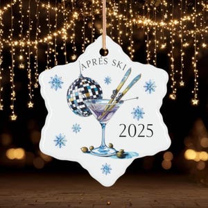 May include: A snowflake-shaped Christmas ornament with an après-ski design. It features a martini glass, skis, a disco ball, and the text "APRÈS SKI 2025". Blue snowflakes and olives complete the design. A gold string is attached for hanging.