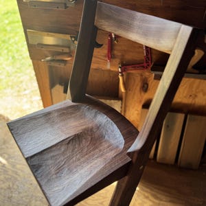 Op de afbeelding: Een donkerbruine houten stoel met een gebeeldhouwde, getextureerde zitting en zichtbare houtnerfpatronen. De stoel heeft een rechte rugleuning en schuine poten, met een natuurlijke afwerking die de diepe tinten van het hout benadrukt.