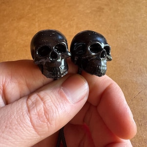 May include: Two small, glossy black skull pendants are held in a hand. The skulls have detailed features, including eye sockets, a nose cavity, and teeth. Each skull is attached to a black cord.