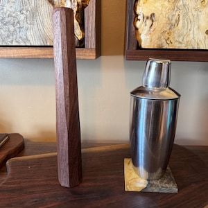 May include: A faceted wooden muddler and a stainless steel cocktail shaker are displayed on a wooden surface. The shaker sits on a small, square coaster. Decorative wooden art pieces are visible in the background.
