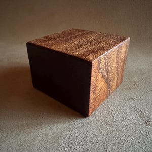 Könnte beinhalten: Ein massiver Holzquader in einem satten, rötlich-braunen Farbton. Die Oberseite zeigt eine ausgeprägte Holzmaserung, während die Seiten eine dunklere, gleichmäßigere Farbe aufweisen. Diese geometrische Form könnte für dekorative oder praktische Zwecke verwendet werden. Maße: ca. 10 cm x 10 cm x 10 cm.