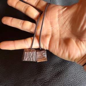 May include: A pair of small, square wooden pendants with carved textures, suspended on a black cord. The pendants are a warm brown color, and the cord appears to be made of a woven material. The background is a dark surface.