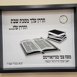 Peut inclure: Une plaque encadrée de noir avec un fond blanc. La plaque présente une illustration en noir et blanc d'un livre, d'une pile de livres et d'une horloge. Le texte sur la plaque est "הדרן עלך מסכת שבת והדרן עלן... משה צבי בערקאוויטש ראש ישיבה תורת חסד המספיק יום א' בשלח תשפ"ה"