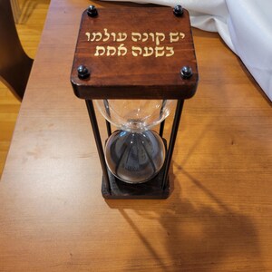 May include: A wooden hourglass with a black metal frame and a glass bulb. The top of the hourglass is engraved with the Hebrew text "Yid Kodesh Shelo Yachliph Be'esh Ha'am."