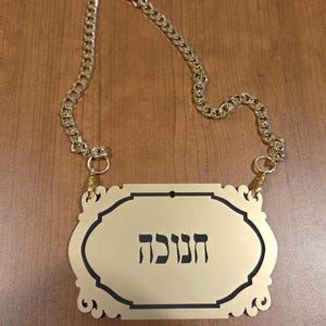 May include: A gold-colored sign with the Hebrew word "Hanukkah" in black lettering. The sign has an ornate, decorative border and is suspended by a gold chain.