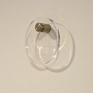 May include: A clear acrylic cabinet knob with a circular design and a brass-colored metal screw. The knob is mounted on a light-colored wall, casting a shadow. This modern hardware is suitable for drawers and cabinets.