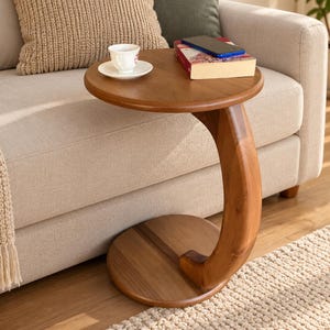 May include: A round wooden side table with a C-shaped base and a single curved support column. The table surface holds a white ceramic teacup on a saucer, a rectangular book, and a blue smartphone. The wood has a warm brown finish.