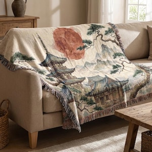 Könnte beinhalten: Beige Überwurfdecke mit japanischem Landschaftsdesign mit einer Pagode, Bergen und einer roten Sonne. Die Decke hat einen Fransenrand und ist über ein beiges Sofa drapiert. Der Überwurf ist ein dekoratives Heimtextil.