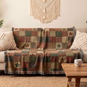 May include: A patchwork throw blanket with star and plaid patterns in shades of brown, green, and red is draped over a beige sofa. Two cream-colored throw pillows and a wooden coffee table complete the cozy living room scene.