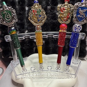 May include: Four decorative pens with jeweled tops and different colored bodies. The pens are in a clear plastic holder with the words "Cute Creations" engraved on the front. The pens feature the crests of the four Hogwarts houses: Gryffindor, Slytherin, Hufflepuff, and Ravenclaw.