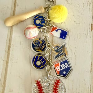 May include: A silver-toned metal keychain featuring a miniature wooden baseball bat, a yellow pom-pom, a small baseball, and several acrylic charms. The charms include the Milwaukee Brewers logo, the MLB logo, and a large glittery baseball disc.