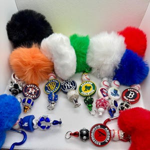 May include: A collection of baseball-themed keychains featuring colorful faux fur pom-poms, team logo charms, and decorative beads. The charms display various Major League Baseball team logos including the Astros, Brewers, Yankees, Athletics, Royals, Red Sox, Dodgers, and Diamondbacks.