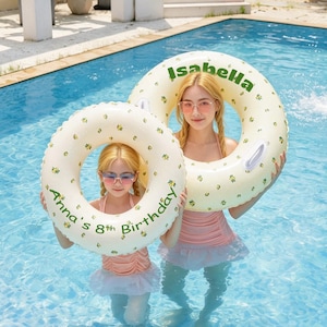 Op de afbeelding: Twee ronde opblaasbare zwembanden van crèmekleurig vinyl met een klein geel bloemenpatroon. Eén zwemband is voorzien van de groene tekst Isabella, de andere van de groene tekst Anna's 8th Birthday in een cirkelvormig lettertype.