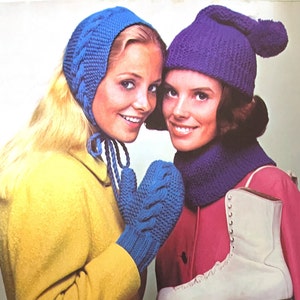 Puede incluir: Dos mujeres con gorros y bufandas de punto. Una mujer lleva guantes azules y un abrigo amarillo. La otra mujer lleva un abrigo rosa y patines de hielo blancos.