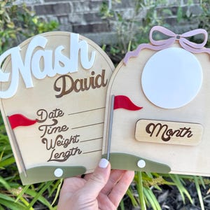 May include: Two wooden baby milestone signs. One sign has the name "Nash David" in white and brown script. The other sign has a circular photo space and the word "Month". Both signs have a red flag and a green base.