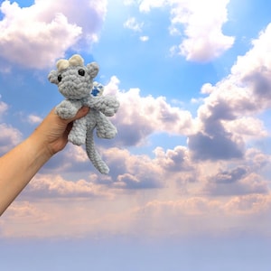Op de afbeelding: Een handgemaakte, lichtgrijze gehaakte draak speelgoed met een lange staart, kleine vleugels en zwarte ogen. De draak wordt omhoog gehouden tegen een blauwe lucht vol pluizige witte wolken. De draak heeft witte accenten op zijn hoofd en een blauwe ster op zijn vleugel.