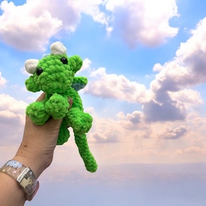 Op de afbeelding: Een handgemaakte groene gehaakte draak met witte hoorns en zwarte ogen. De draak wordt omhoog gehouden tegen een blauwe lucht met pluizige witte wolken. De draak heeft een lange staart en kleine vleugels.