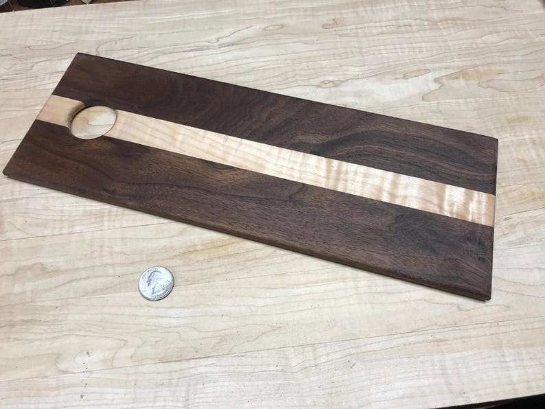 Op de afbeelding: Een rechthoekig houten serveerplank met een donkerbruine walnoot buitenkant en een lichtgekleurde esdoorn streep in het midden. Een cirkelvormig gat is in &eacute;&eacute;n uiteinde uitgesneden. Een kwart dollar munt ligt naast de plank.