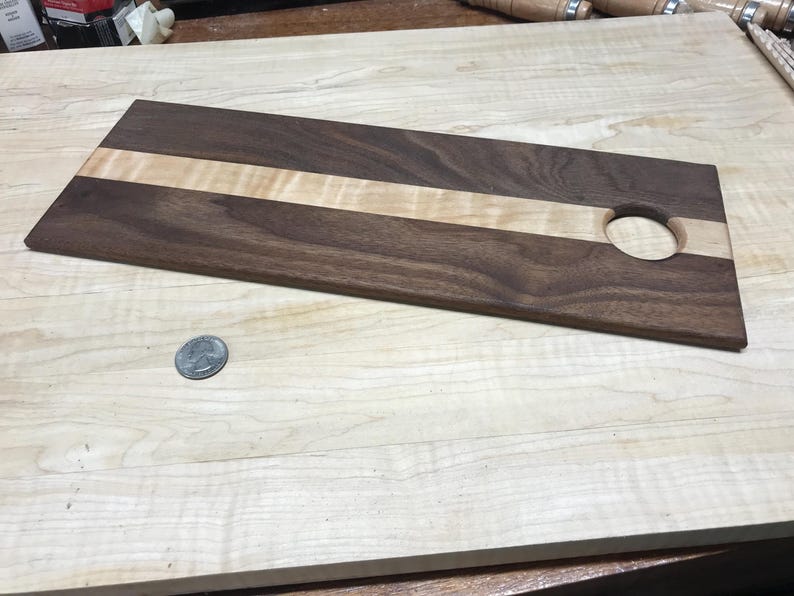 Op de afbeelding: Een rechthoekig houten serveerplank met een donkerbruine walnoot buitenkant en een lichtgekleurde esdoorn streep in het midden. Het heeft een cirkelvormig gat aan &eacute;&eacute;n uiteinde. De plank is ongeveer 40 cm lang.