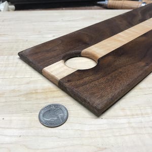 Op de afbeelding: Een rechthoekige houten serveerplank met een donkerbruine afwerking en een lichtgekleurde streep in het midden. De plank heeft een cirkelvormige uitsparing en is ongeveer 30 cm lang. Een munt ligt naast de plank ter referentie.