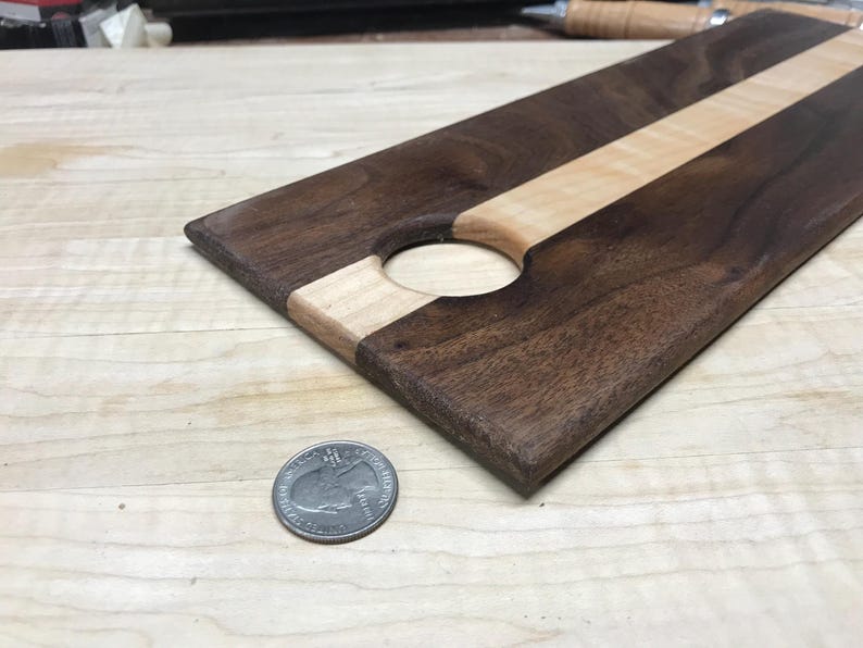 Op de afbeelding: Een rechthoekige houten serveerplank met een donkerbruine afwerking en een lichtgekleurde streep in het midden. De plank heeft een cirkelvormige uitsparing en is ongeveer 30 cm lang. Een munt ligt naast de plank ter referentie.