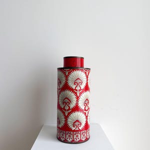 May include: A cylindrical container with a red background and a repeating white floral pattern. The container has a red lid and base. The design is symmetrical and the container is placed on a white surface.