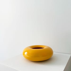 Op de afbeelding: Een glanzende, donutvormige keramische vaas in een levendige gele kleur. De binnenkant is diep roodbruin. De vaas staat op een wit oppervlak, wat een opvallend contrast creëert. Dit decoratieve stuk voegt een vleugje kleur en moderne flair toe aan elke ruimte.
