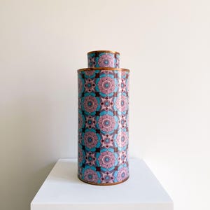 May include: A cylindrical metal canister featuring a repeating geometric floral pattern in shades of pink, light blue, and dark brown. The container has a copper-toned rim at the top and a matching copper-toned base, with a smaller cylindrical lid.