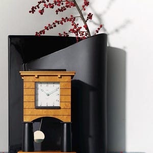 Puede incluir: Un reloj de madera con esfera cuadrada y péndulo, colocado frente a un jarrón negro y una ramita de bayas rojas. El reloj tiene un acabado de madera clara con detalles en negro y una esfera blanca.