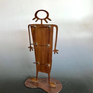 以下が含まれることがあります： 円形の頭とシンプルなデザインの人物を描いた、さび付いた金属製の彫刻。彫刻は平らな土台の上に立っており、暗い表面の上に置かれています。