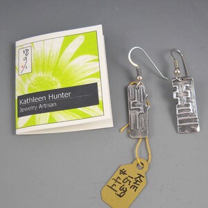 May include: A pair of silver earrings with a geometric design. The earrings are hanging from a white card with the text "Kathleen Hunter Jewelry Artisan".