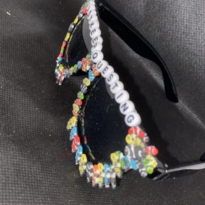 May include: Black sunglasses with a colorful beaded frame that spells out "Keep Questioning".
