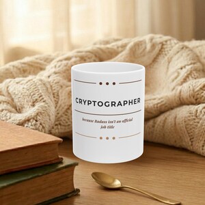 May include: White ceramic mug with the word "CRYPTOGRAPHER" in a brown font, with the phrase "because Badass isn't an official job title". The mug is on a wooden surface next to two books and a gold spoon.