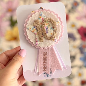 May include: A handmade birthday badge in pink and white. The badge has a star and the number "10" in gold glitter, with the name "Hermione" on a pink ribbon. The badge is on a white card. The background is floral.
